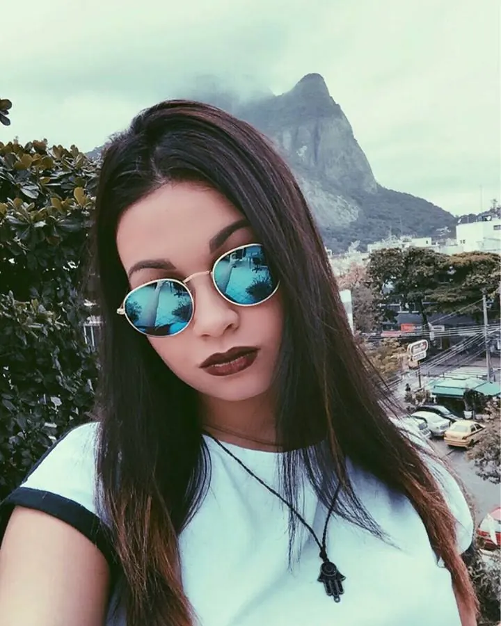 Femme portant des lunettes de soleil rondes, avec un collier de main, devant un paysage urbain.