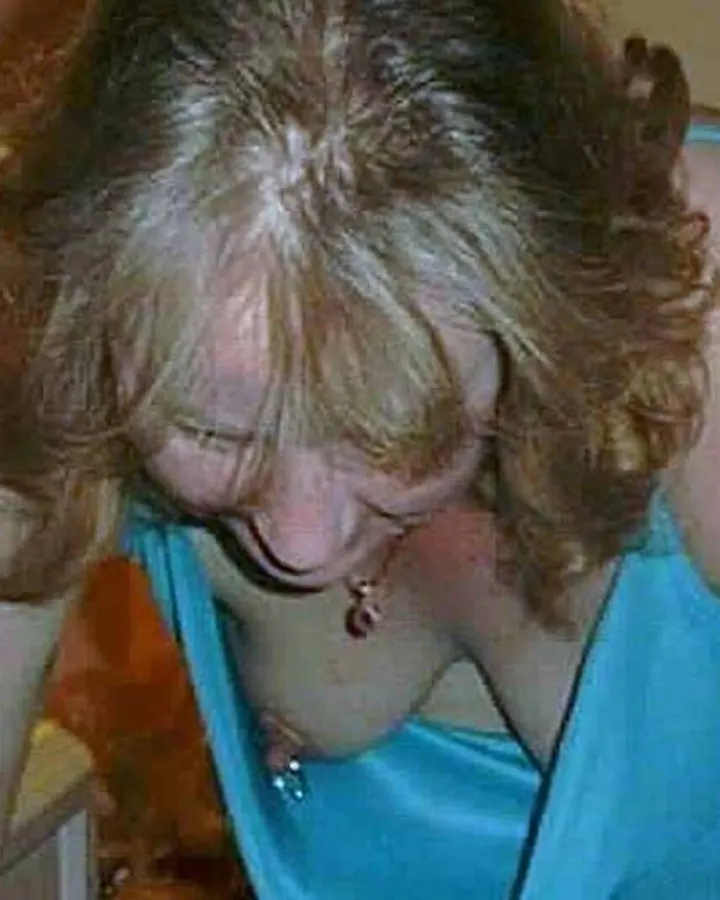 Femme blonde penchée en avant, portant un haut bleu, dans une pièce éclairée par une lampe.
