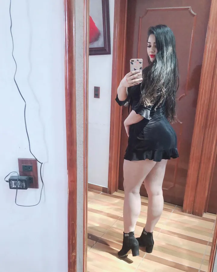 Femme aux cheveux noirs, vêtue d'une chemise transparente blanche, posant contre un mur blanc, tatouages visibles, regard direct.