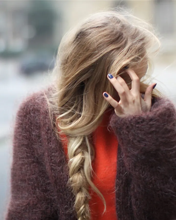 Femme aux cheveux blonds, vêtue d'un pull orange et d'une veste marron, marchant dans la rue, main devant le visage, regard caché.