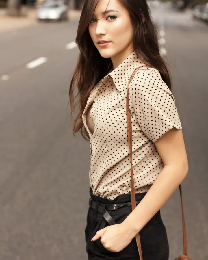 Femme aux cheveux châtains et yeux marrons, marchant dans la rue, portant une chemise à pois et une jupe noire.