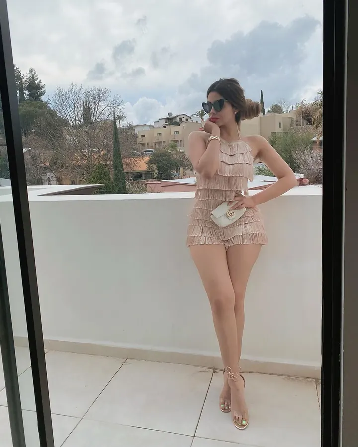 Femme aux cheveux châtains et yeux marrons, posant dans une rue, portant une robe à franges beige et des lunettes de soleil.