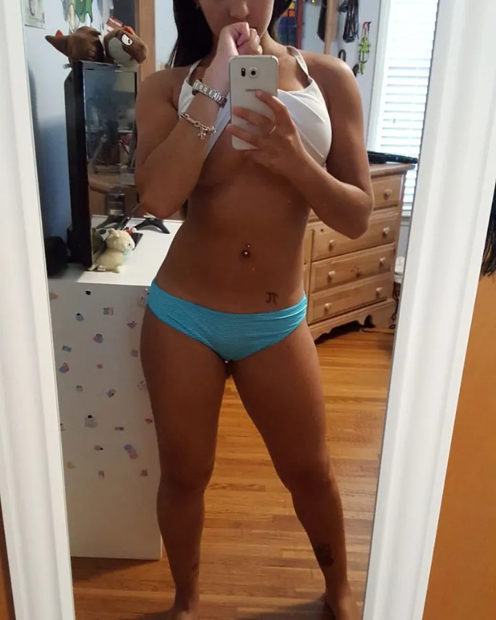 Jeune femme aux cheveux bruns-noirs et yeux noirs, prenant un selfie devant un miroir, portant un haut blanc et un short bleu.