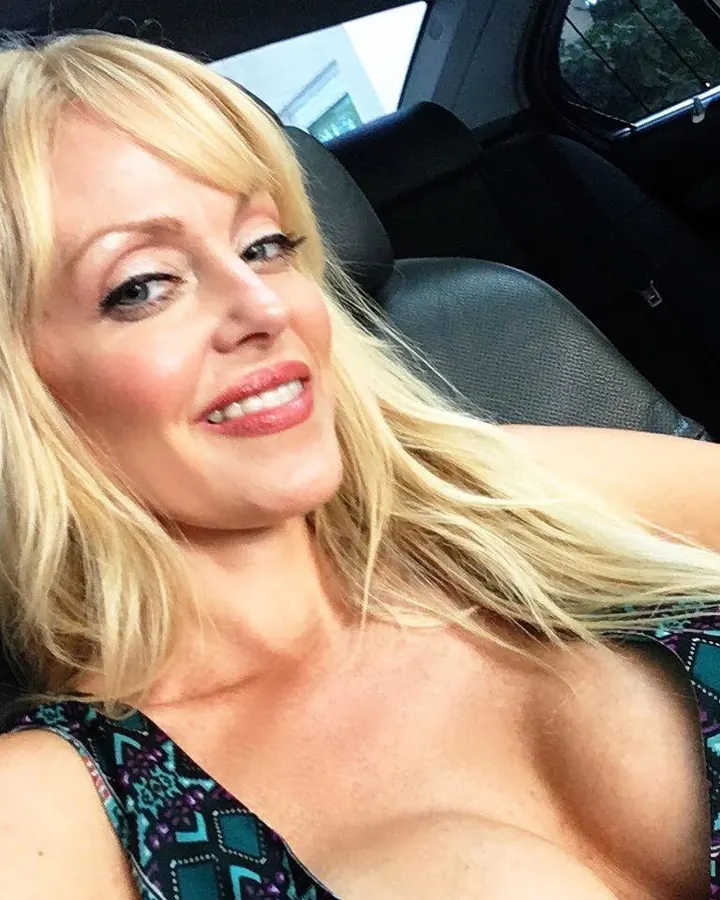 Femme blonde aux yeux bleus, souriante, allongée sur un siège de voiture, vêtue d'un haut coloré.
