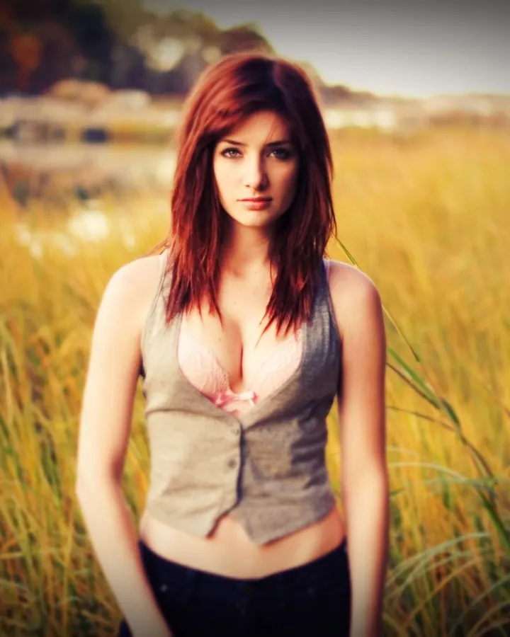 Femme européenne aux cheveux roux, en haut gris et soutien-gorge rose, debout dans un champ doré.