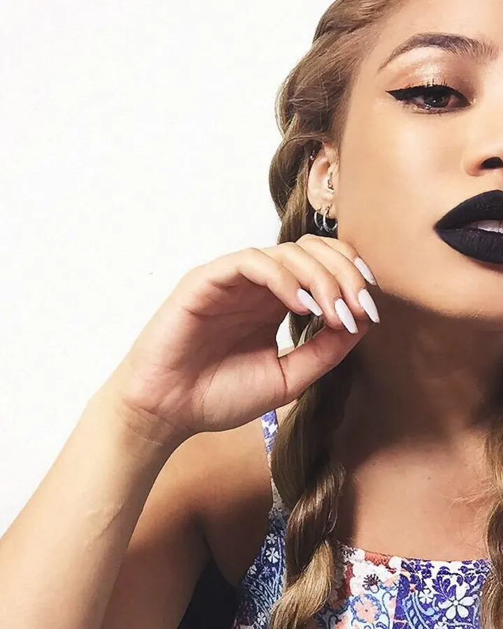 Femme aux cheveux blonds, maquillée avec du rouge à lèvres noir, regardant vers le côté, la main posée sur sa joue, avec des ongles blancs.