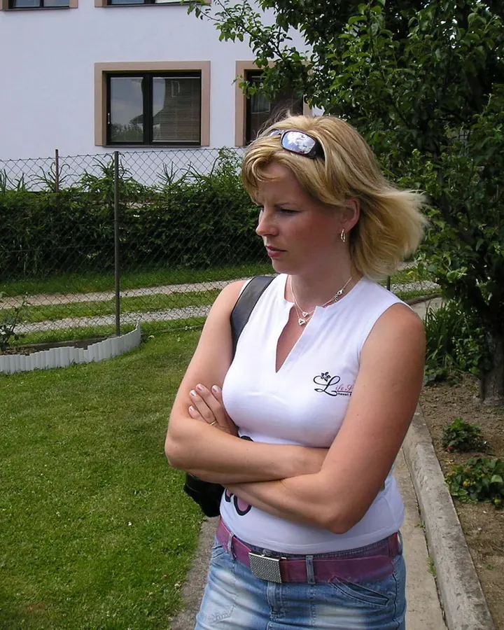 Femme aux cheveux blonds, en tenue décontractée, portant un haut blanc et un jean, debout dans un jardin, les bras croisés.
