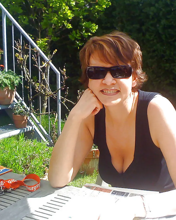 Femme d'âge mûr en débardeur noir, assise à une table de jardin, portant des lunettes de soleil, souriant légèrement.