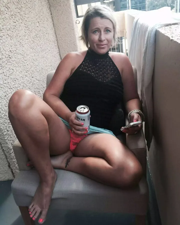 Femme blonde assise sur un fauteuil, tenant une canette, en tenue noire et rose.