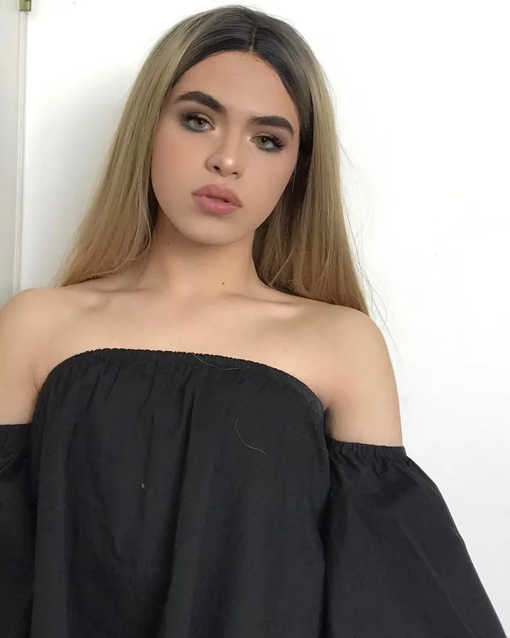 Femme blonde en haut noir décolleté, posant contre un mur blanc