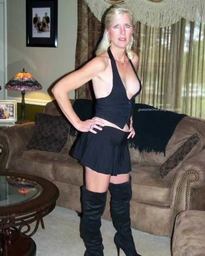 Femme aux cheveux blonds, posant debout dans un salon, portant une robe noire profondément décolletée et des bottes noires à talons.