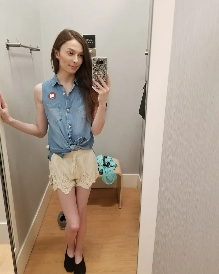 Jeune femme aux cheveux châtains, prenant un selfie dans un vestiaire, portant un haut en jean sans manches et un short beige.