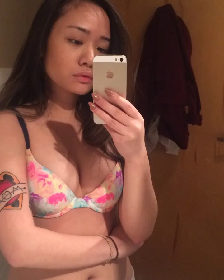 Jeune femme aux cheveux bruns noirs, prenant un selfie dans un miroir, portant un soutien-gorge floral coloré, tatouage visible sur le bras.