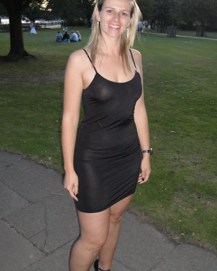 Femme aux cheveux blancs, souriante, portant une robe noire ajustée, debout sur un trottoir dans un parc.