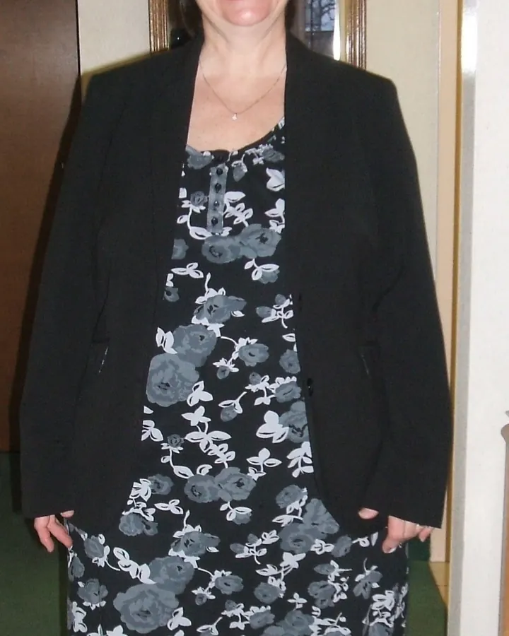 Femme aux cheveux bruns courts, souriante, portant une robe noire à imprimé floral, une veste noire, des collants noirs et des chaussures noires, debout dans un couloir.