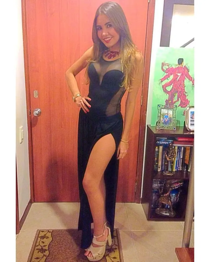 Femme aux cheveux bruns longs, souriante, portant un haut blanc à découpes, un short noir, une bague, une perle à l'oreille, prenant un selfie dans une chambre.
