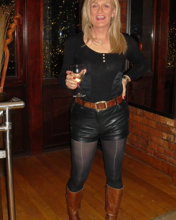 Femme blonde souriante, tenant un verre de vin, vêtue d'un haut noir, d'un short en cuir noir, de collants noirs et de bottes marron, debout sur un sol en bois.