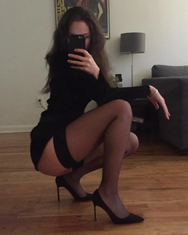 Femme aux cheveux bruns, en robe noire courte, agenouillée sur un parquet, tenant son téléphone devant son visage, chaussée de talons noirs.