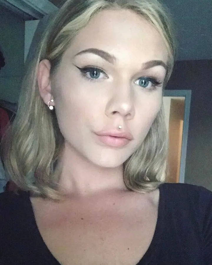 Portrait d'une jeune femme blonde aux yeux bleus, maquillée, avec un piercing au nez, portant un haut noir.