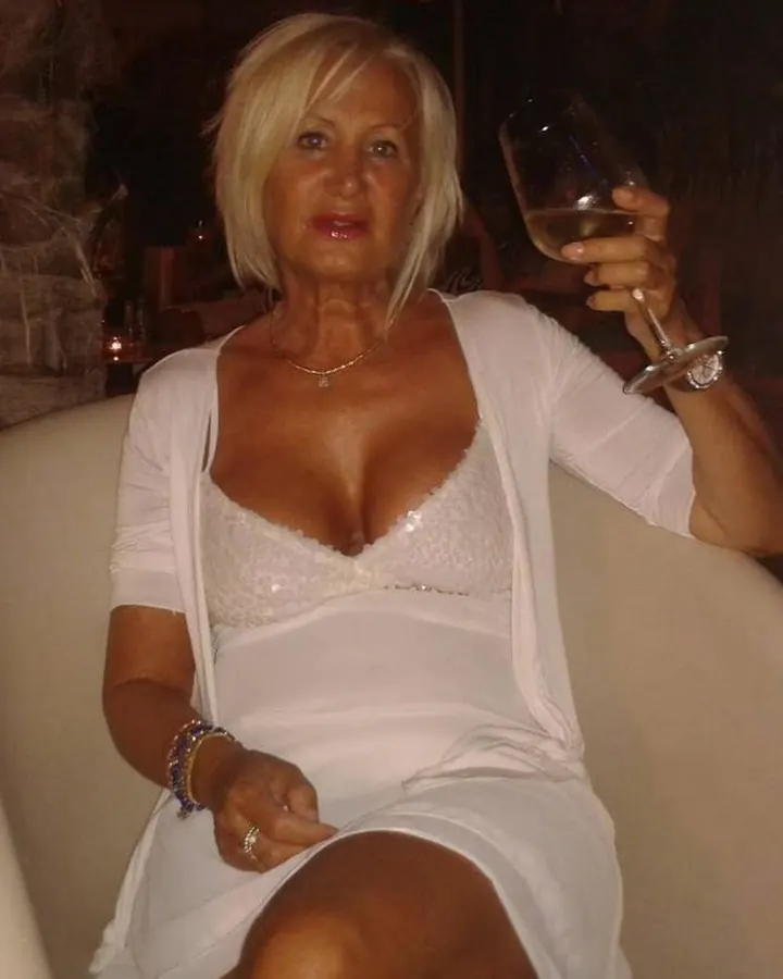 Femme blonde assise, portant une robe blanche et un cardigan, tenant un verre de vin blanc dans un cadre sombre.