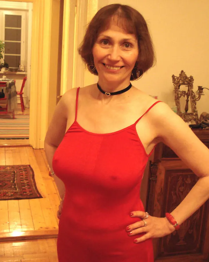 Femme aux cheveux châtains courts, en robe rouge ajustée, souriante, debout dans un salon avec un meuble ancien en bois derrière elle.