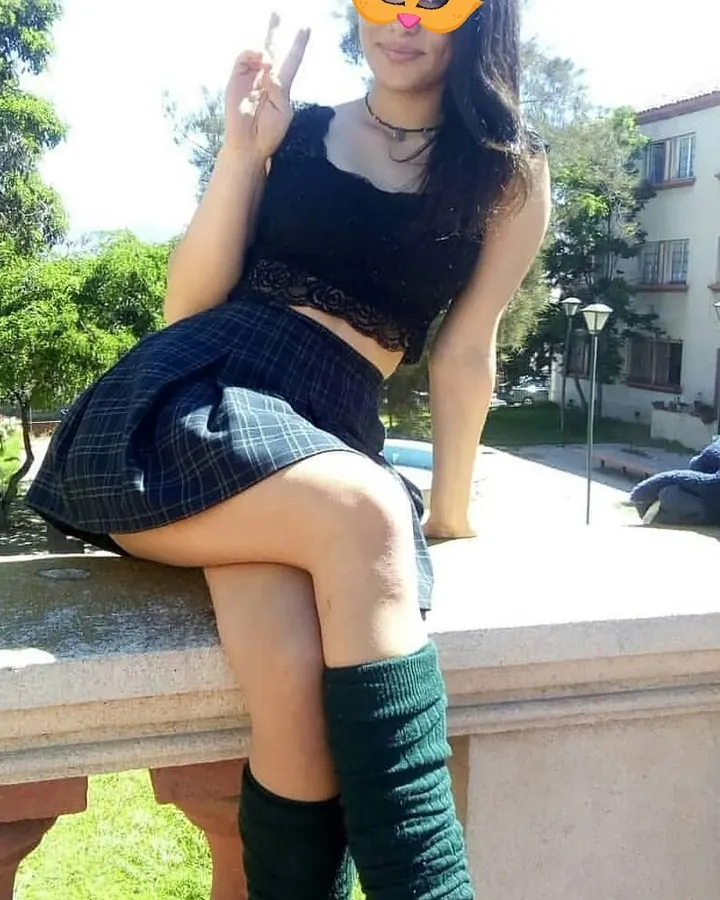 Jeune femme aux cheveux noirs longs, en tenue scolaire (débardeur noir, jupe à carreaux), assise sur un banc, les genoux couverts de collants verts, masque orange sur le visage.