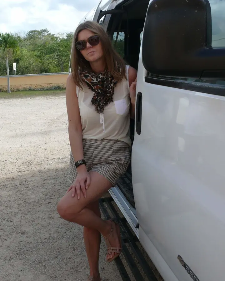 Femme brune en tenue décontractée, assise sur le seuil d'un véhicule blanc, portant des lunettes de soleil et un foulard léopard.