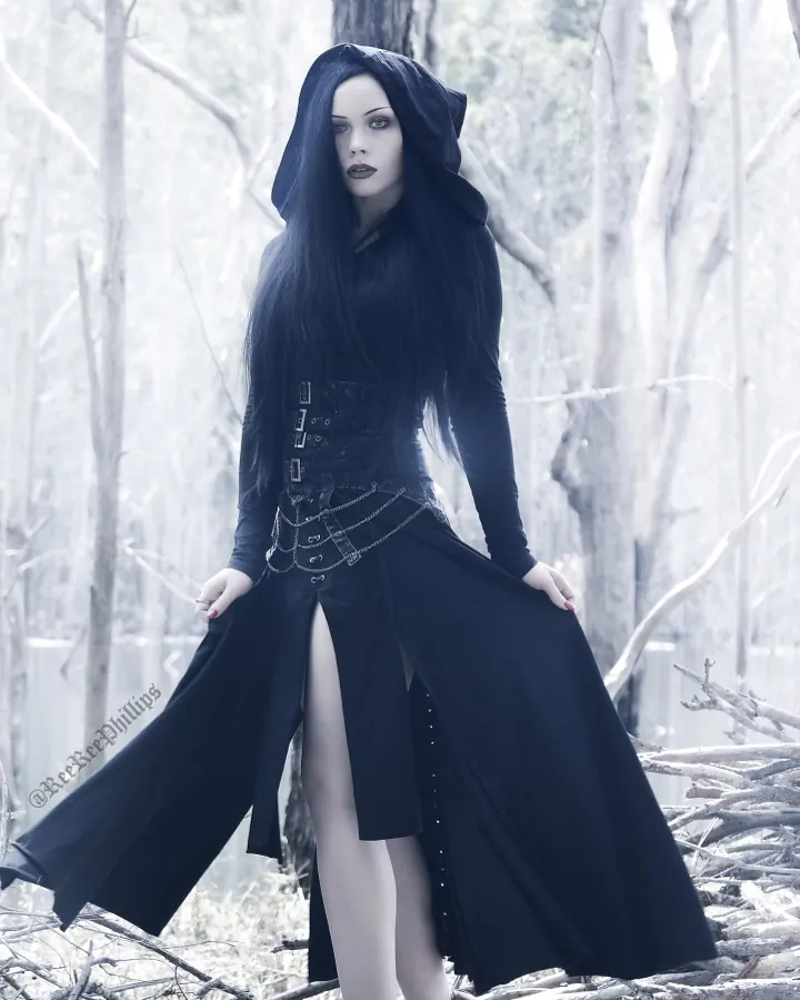 Femme aux cheveux noirs, portant un costume gothique noir avec capuche, debout dans une forêt morte, tenant sa robe ouverte.