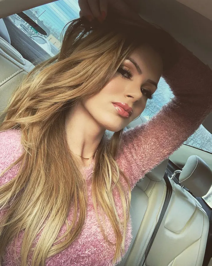 Femme blonde en pull rose, assise dans une voiture, les cheveux tombant sur son visage, regardant vers le bas.