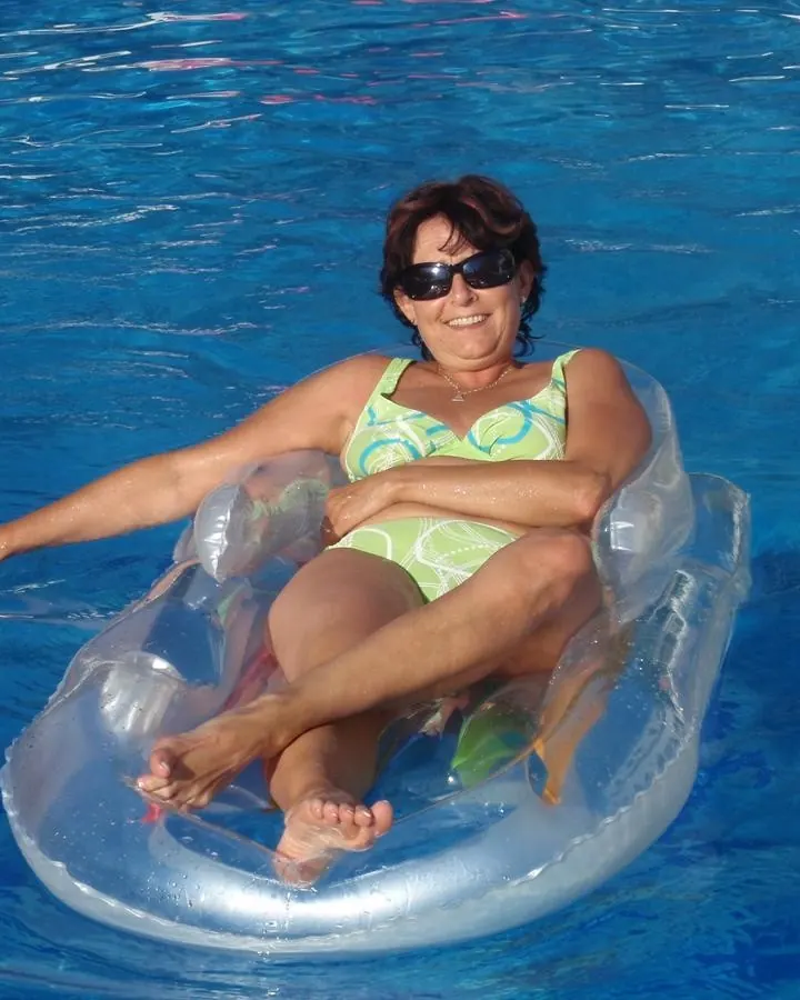 Femme aux cheveux bruns, flottant sur un matelas pneumatique dans une piscine bleue, portant un maillot de bain vert et jaune.