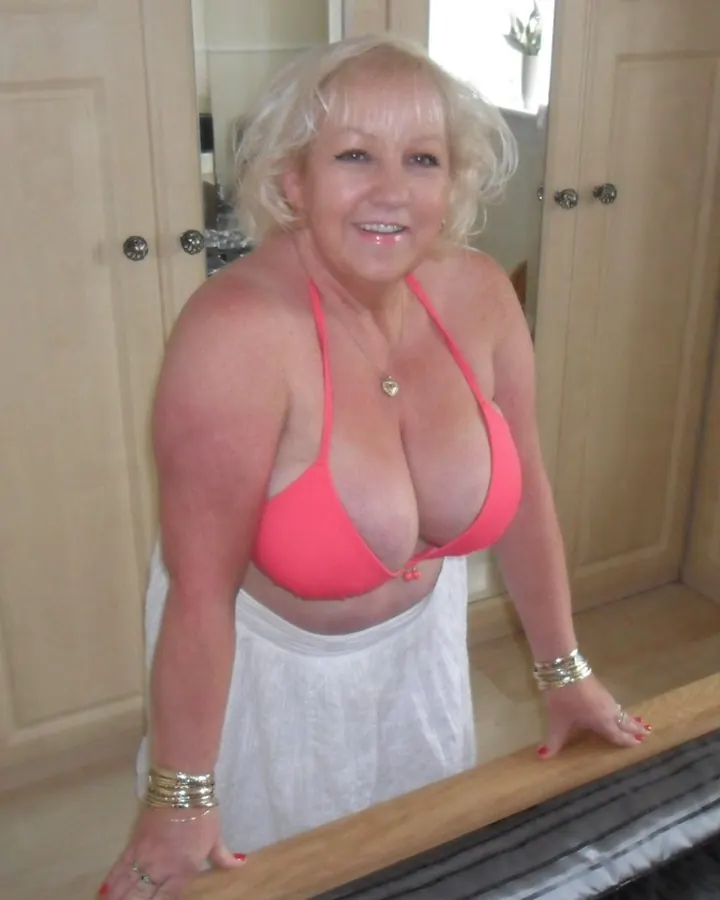 Femme blonde en soutien-gorge rose, souriante, appuyée sur un lit dans une chambre.