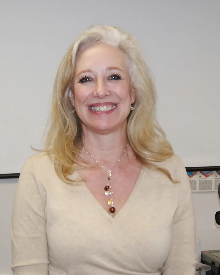 Femme aux cheveux blonds souriant à la caméra en chemisier beige, dans une salle de classe.