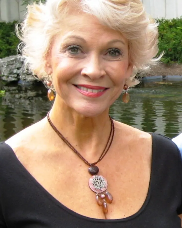 Femme aux cheveux blonds courts, souriant, portant un collier et une robe noire, devant un étang.