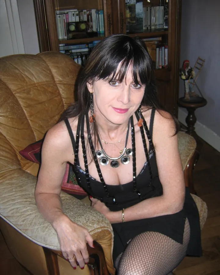 Femme aux cheveux bruns-noirs assise sur un fauteuil en cuir, portant une robe noire et des collants résille.