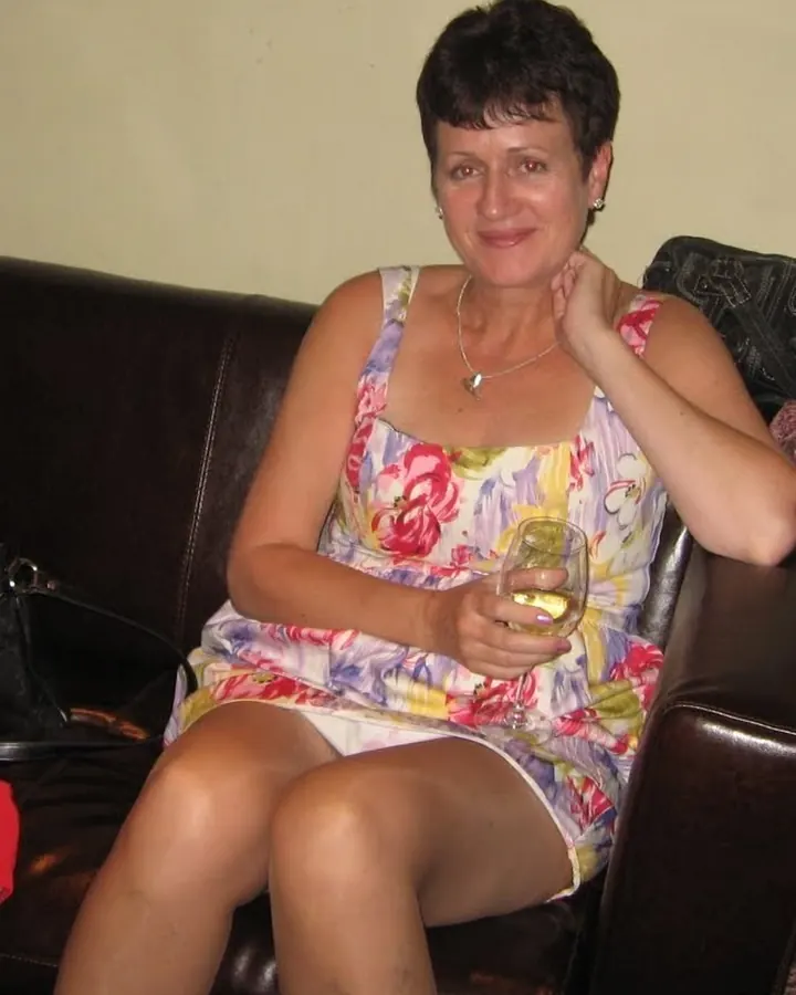 Femme européenne aux cheveux courts, en robe fleurie, assise sur un canapé noir, tenant un verre de vin blanc.