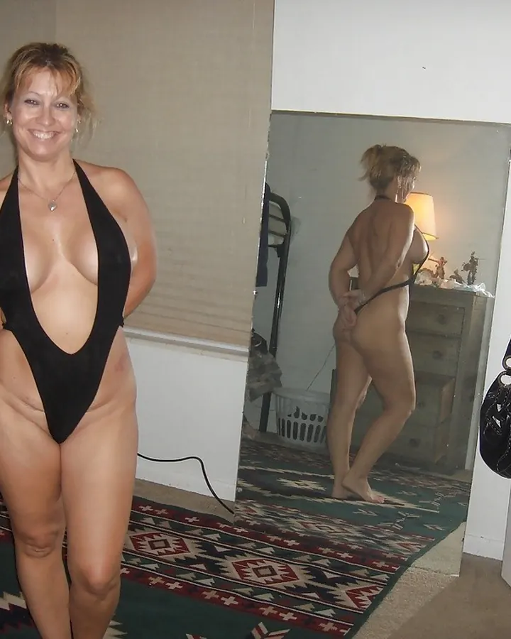 Femme européenne aux cheveux blonds, en robe noire, posant sur un lit, avec des collants et des chaussures noires.