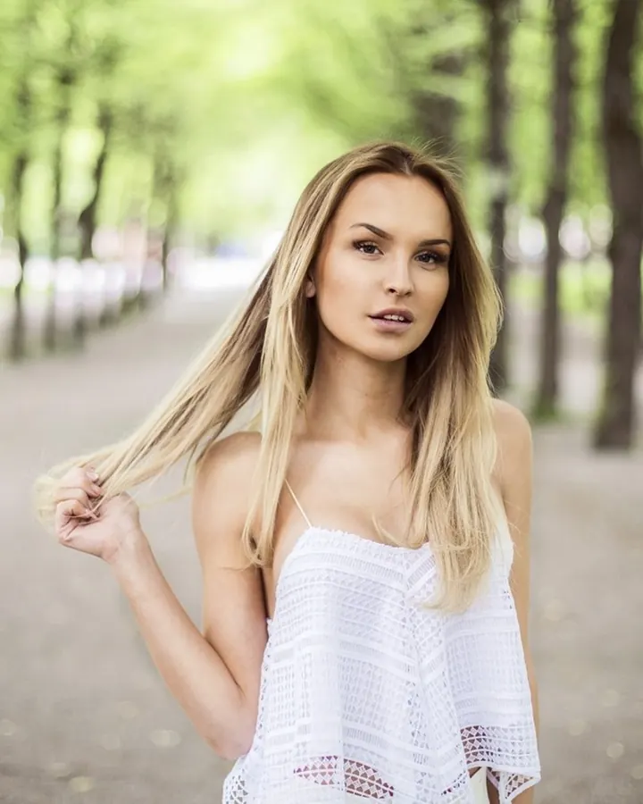 Femme blonde, souriante, portant un haut blanc, posant sur un lit blanc.
