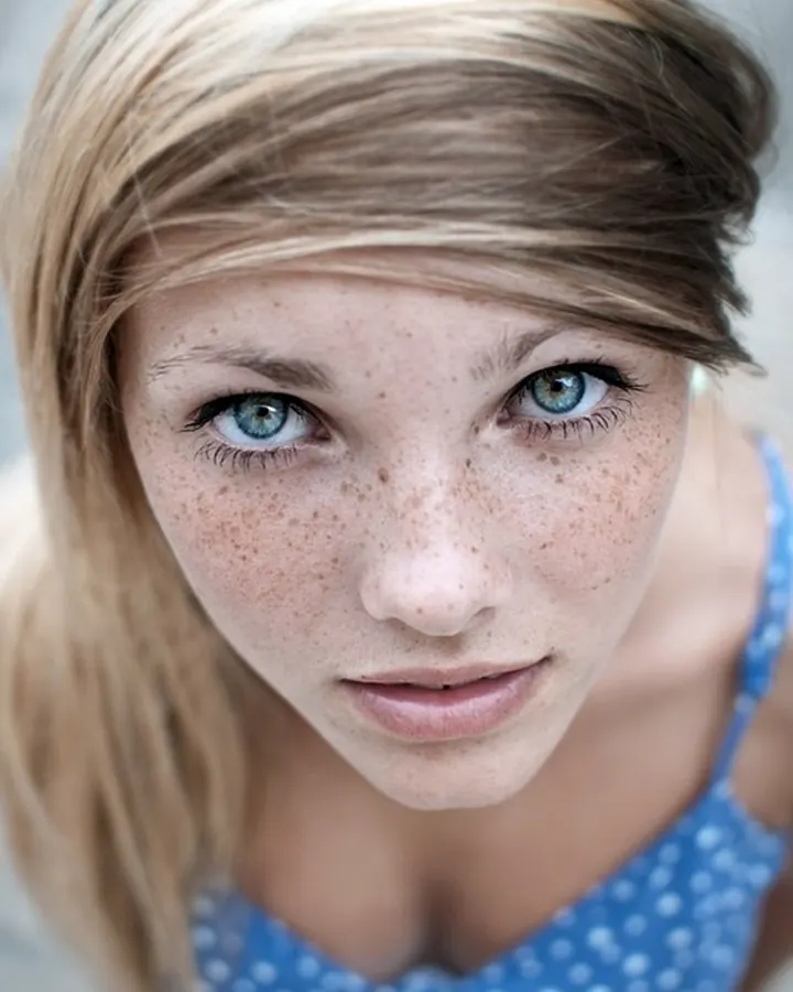 Femme blonde, souriante, portant un haut bleu, posant sur un lit blanc.