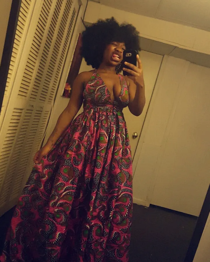 Femme aux cheveux afro noirs, portant une robe longue à motifs colorés, prenant un selfie devant un miroir avec une expression joyeuse.