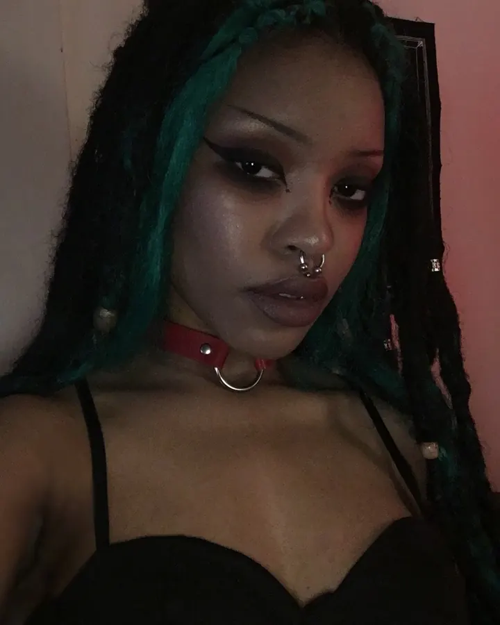 Femme aux cheveux noirs et verts, portant un collier rouge avec anneau, un haut noir et un piercing nasal, regardant directement l'appareil photo dans une pièce sombre.