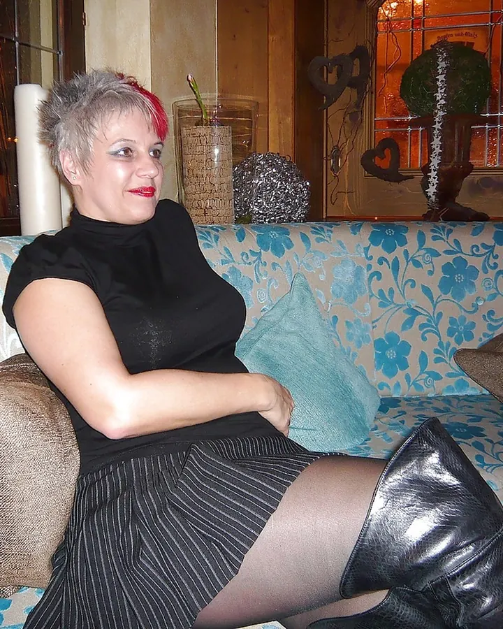 Femme aux cheveux gris avec une mèche rouge, assise sur un canapé à motifs floraux, portant une robe noire, des collants et des bottes en cuir noir.