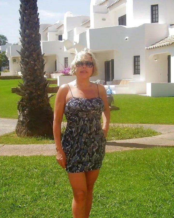 Femme blonde, debout dans un jardin, portant une robe courte à motifs bleus et noirs, avec des lunettes de soleil, devant des bâtiments blancs et une pelouse verte.