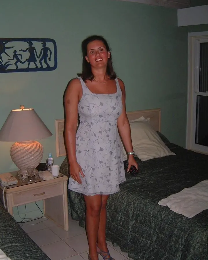 Femme brune debout dans une chambre, robe grise à motifs, tenant une télécommande