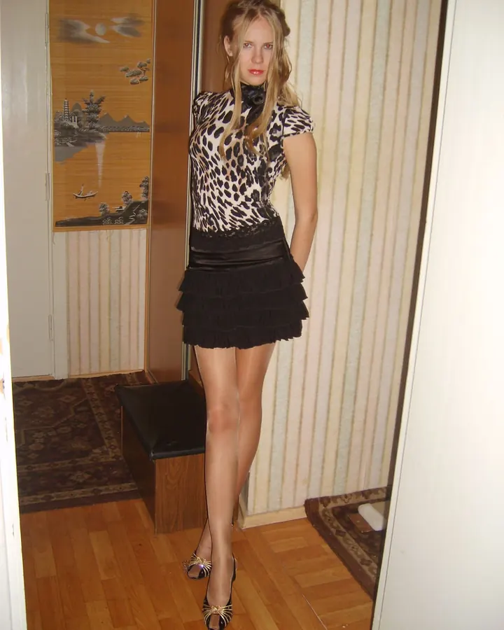 Femme aux cheveux blonds, portant un haut rouge et un short beige, assise sur une commode en bois dans une chambre.