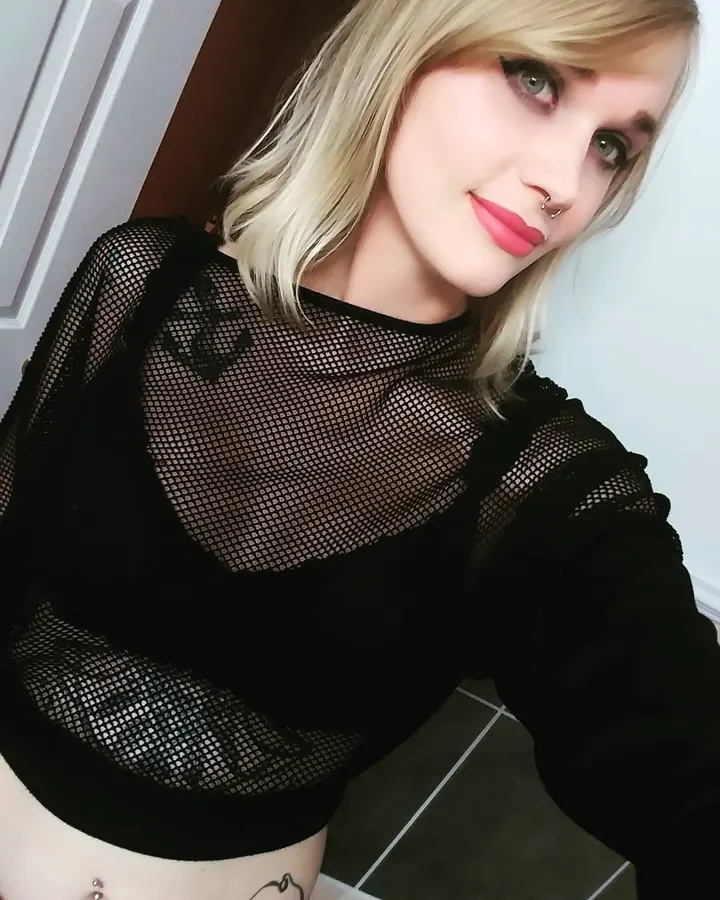 Femme aux cheveux blonds courts et yeux bleus, portant un haut noir transparent à mailles, prend un selfie dans une pièce intérieure.