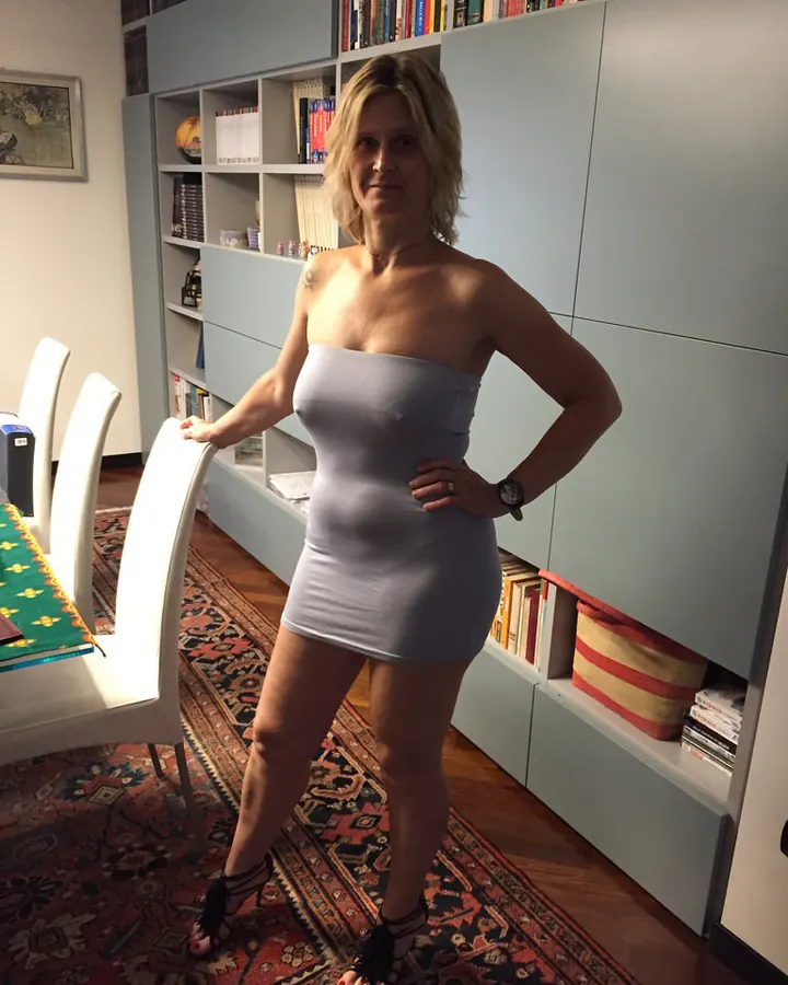 Femme aux cheveux blonds, portant une robe bustier grise, pose debout dans un salon avec une bibliothèque en arrière-plan.