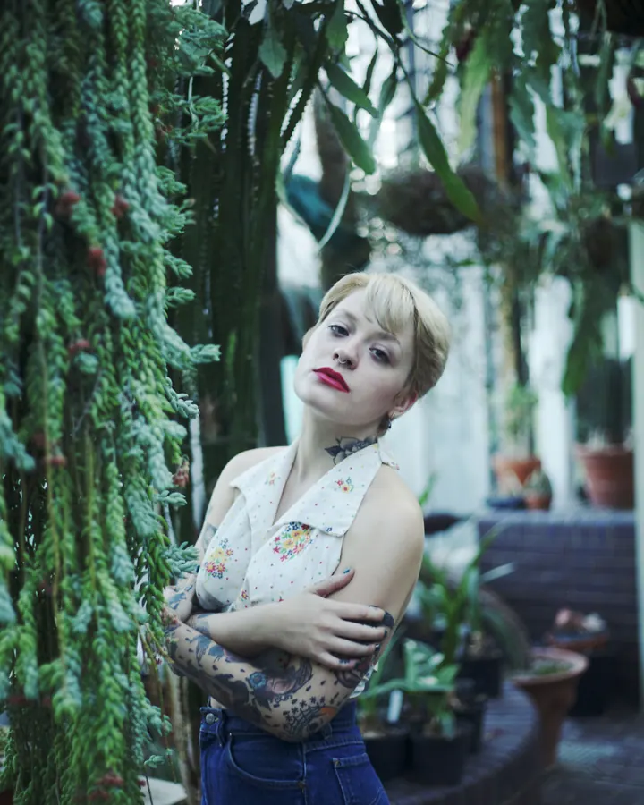Femme aux cheveux châtains, portant une robe noire sans manches, debout contre un arbre dans un jardin, avec des bottes en cuir noir et un tatouage floral sur la jambe.