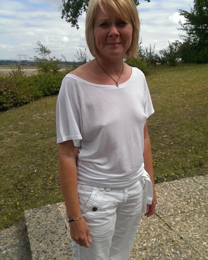 Femme blonde aux cheveux courts, portant une tenue blanche, debout sur un trottoir en pierre, dans un champ verdoyant avec des arbres en arrière-plan.