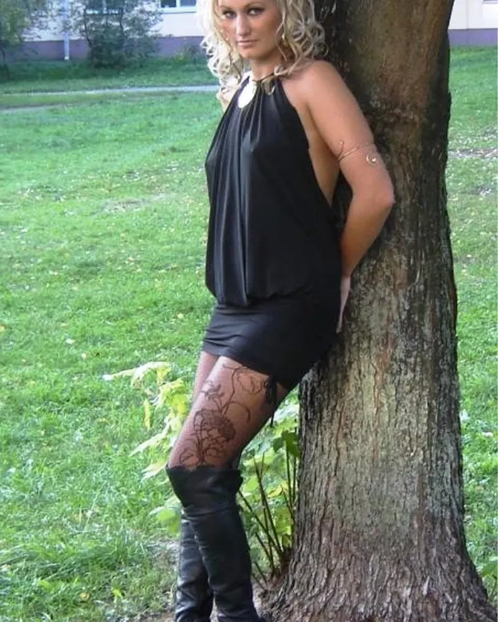 Femme aux cheveux châtains, portant une robe noire sans manches, debout contre un arbre dans un jardin, avec des bottes en cuir noir et un tatouage floral sur la jambe.