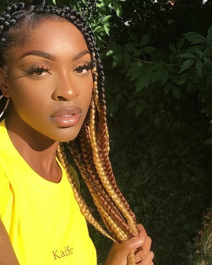 Femme africaine aux cheveux bruns noirs, portant un t-shirt jaune vif, avec des tresses colorées, tenant une mèche de cheveux, devant un arrière-plan végétal.